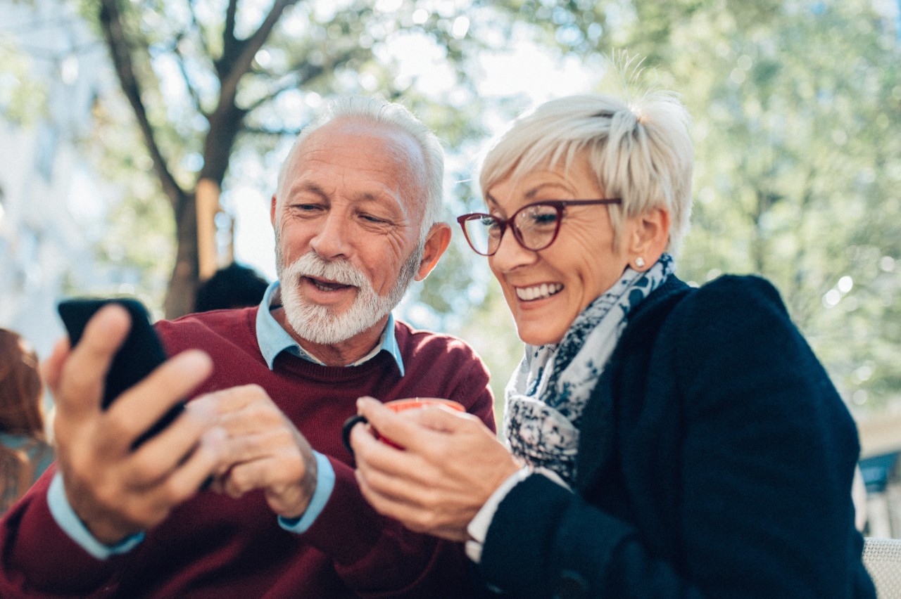 Modern Mature couple using smartphone sitting outdoor in sidewalk cafe in sunny day